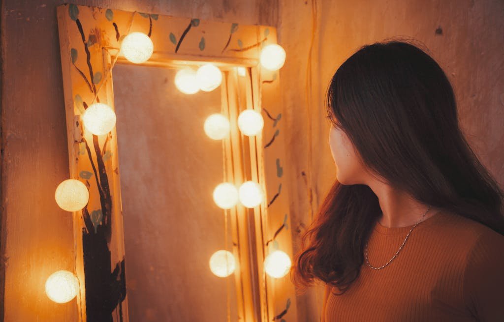 Woman gazing into a warmly lit mirror surrounded by decorative bulbs.